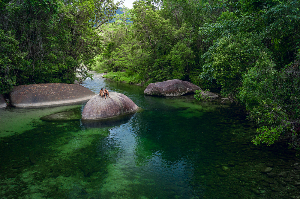 Two people sitting on a large rock in the clear green waters of Babinda Boulders, surrounded by dense rainforest