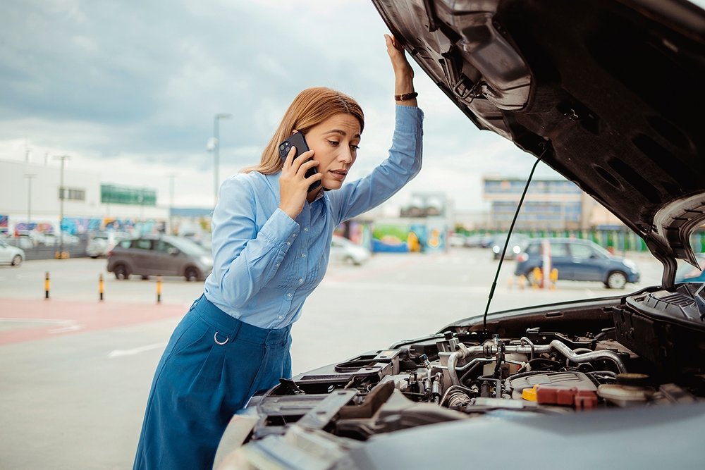 woman calling roadside assistance while looking at car engine