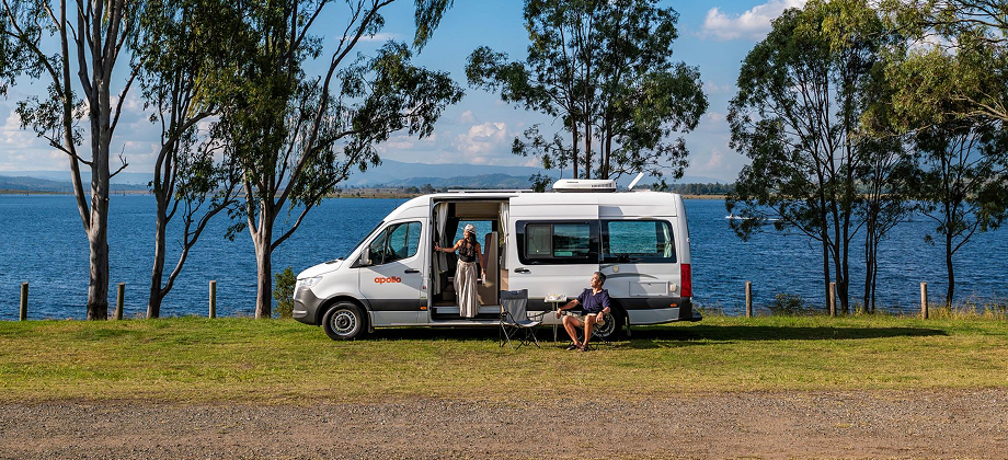 Campervan parked by a lakeside with two people relaxing outdoors under trees.